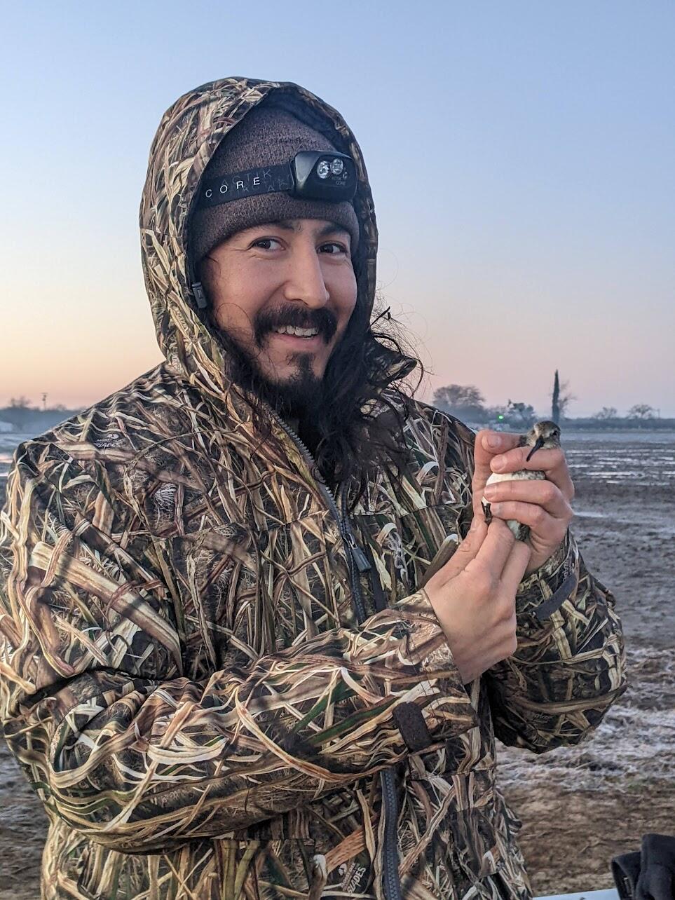 Xerónimo Castañeda holds a Dunlin in his hands while working on a Central Valley farm. The background features a light pink and blue sky just before sunrise, creating a serene and atmospheric setting.
